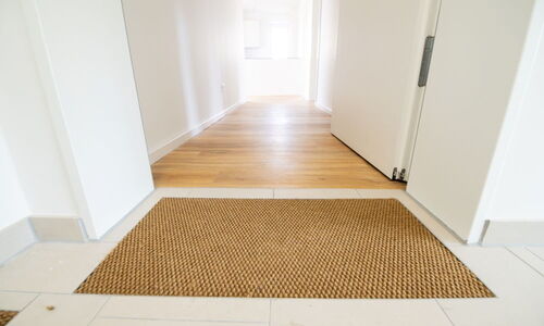 Eingang mit hellbraunem Fußabtreter auf weißem Fliesenboden, offene Tür führt zu Flur mit Holzboden