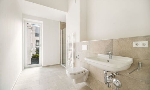 Helles Badezimmer mit Wand-WC, Waschbecken, Handtuchheizkörper und bodentiefem Fenster mit Blick auf Nachbargebäude