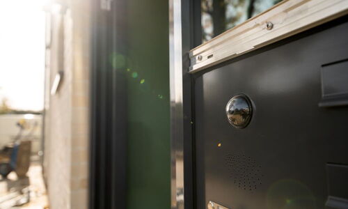 Türsprechanlage mit Kamera und Lautsprecher an einem Gebäude, unscharfer Hintergrund mit Sonnenlicht