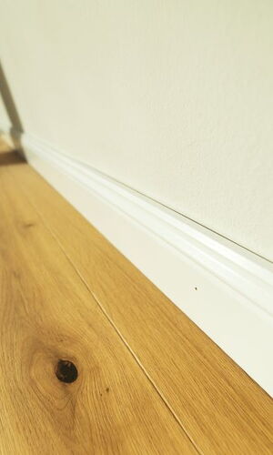 Nahaufnahme eines hellen Holzbodens mit sichtbarer Maserung und weißer Sockelleiste an einer Wand